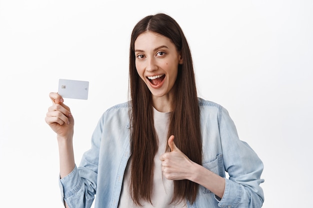 L'immagine della studentessa dice di sì, mostra la carta di credito e il pollice in su, approva e consiglia la banca, un facile pagamento senza contatto, in piedi sul muro bianco