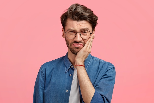 L'immagine di un giovane ragazzo barbuto dispiaciuto ha un'espressione facciale cupa, tocca la guancia con la mano, vestito con una camicia di jeans alla moda, posa contro il muro rosa, essendo insoddisfatto di qualcosa