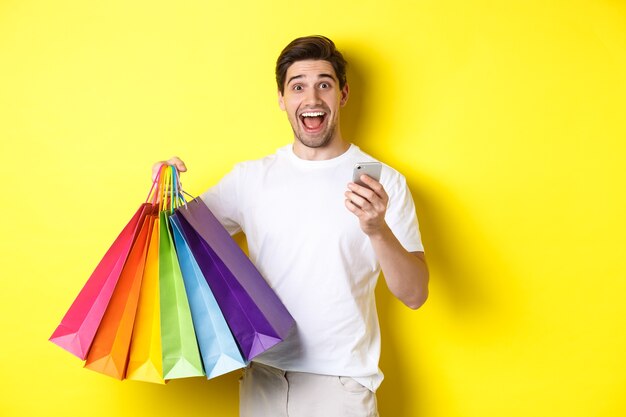 L'immagine di un uomo felice riceve il cashback per l'acquisto, tenendo in mano lo smartphone e le borse della spesa, sorridendo eccitato, in piedi su sfondo giallo.