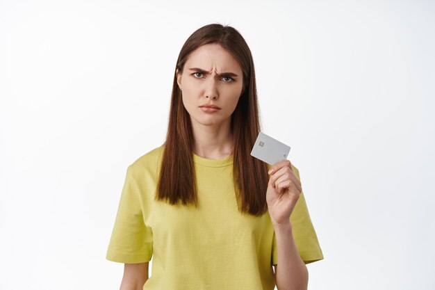 L'immagine di una donna accigliata sembra scontrosa, mostra la carta di credito e sembra arrabbiata per qualcosa, non ha soldi sul conto bancario, problemi con il pagamento, in piedi su sfondo bianco.