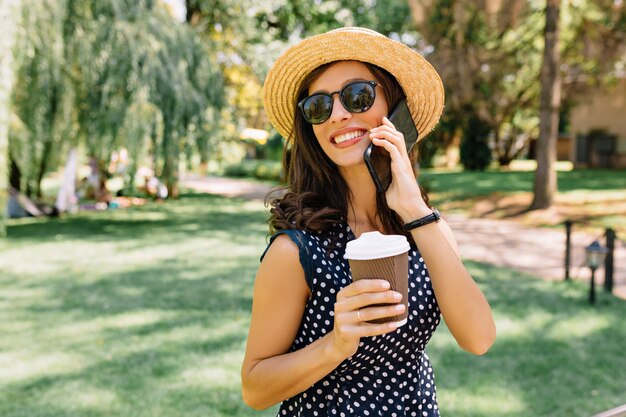 L'immagine di una donna dallo stile affascinante sta camminando nel parco estivo indossando un cappello estivo e occhiali da sole neri e un vestito carino. Beve caffè e parla al telefono con grandi emozioni.