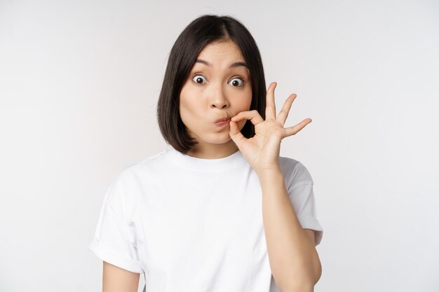L'immagine di una giovane donna asiatica che mostra il gesto di silenzio chiudendo la bocca con le labbra del sigillo fa una promessa in piedi su uno sfondo bianco