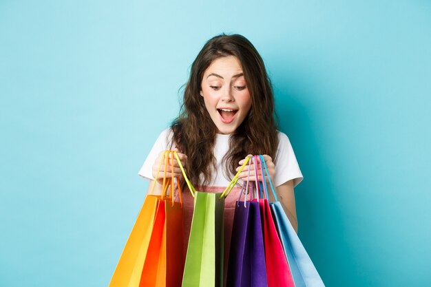 L'immagine di una giovane donna felice porta molte borse della spesa, compra cose su sconti primaverili, in piedi su sfondo blu.
