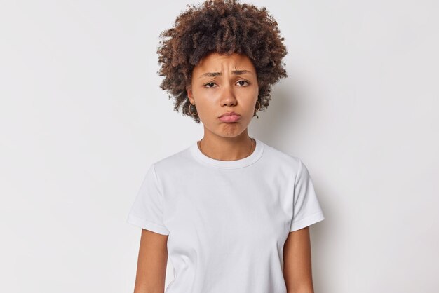 L'immagine di una giovane donna infelice con i capelli ricci si sente disgustosa o di pietà fa smorfie dispiaciute aggrotta le sopracciglia sconvolta vestita con una maglietta casual isolata su sfondo bianco pensa a qualcosa di fastidioso