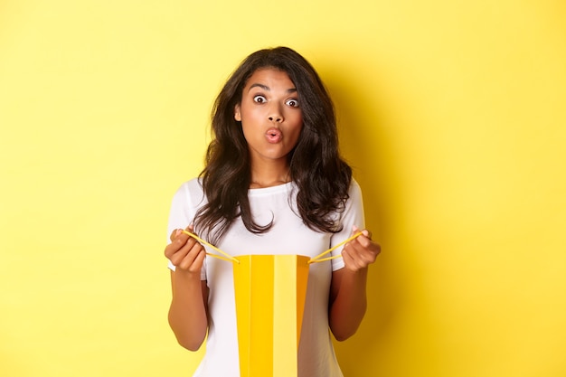 L'immagine di una ragazza afroamericana sorpresa riceve un regalo in vacanza, apre una borsa della spesa e sembra stupita, in piedi su sfondo giallo.