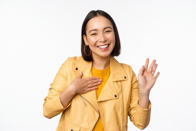 L'immagine di una ragazza asiatica amichevole in un elegante braccio di sollevamento del cappotto giallo si presenta salutando la mano agitando dicendo ciao in piedi su sfondo bianco