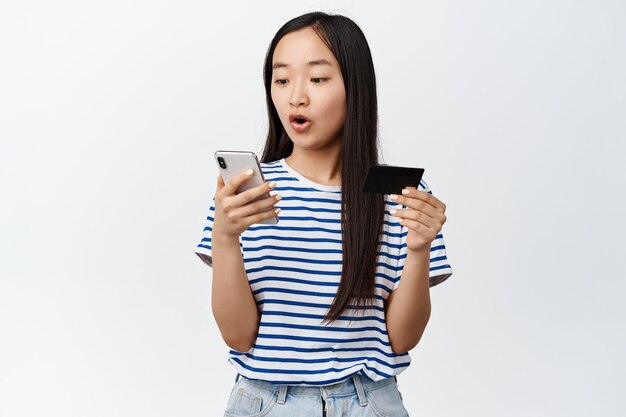 L'immagine di una ragazza asiatica in possesso di carta di credito, sembra entusiasta dello schermo del telefono cellulare, paga per lo shopping online, utilizza l'app del negozio, in piedi su bianco.