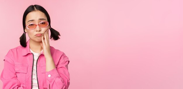 L'immagine di una ragazza asiatica triste che tiene il broncio toccando la sua guancia facendo il broncio deluso ha mal di denti in piedi su sfondo rosa