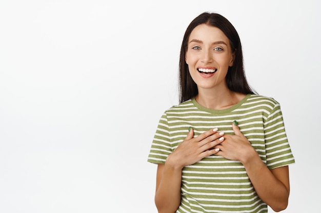 L'immagine di una ragazza bruna soddisfatta e grata guarda con apprezzamento tenendosi per mano sul petto in piedi in maglietta su sfondo bianco