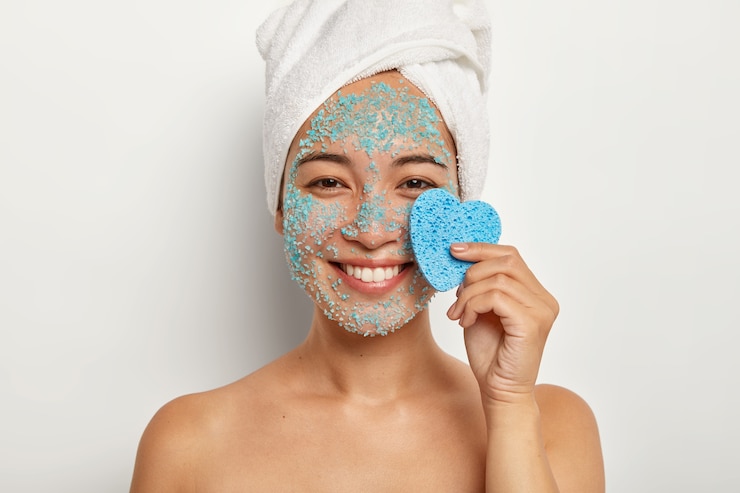 Foto gratuito l'immagine ritagliata di una giovane donna felice tiene una spugna cosmetica a forma di cuore, applica una maschera blu naturale, ha un ampio sorriso, un asciugamano avvolto sulla testa, sta spalle nude al coperto. concetto di trattamenti termali