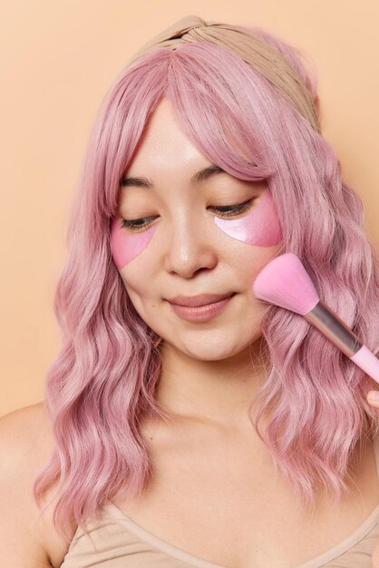 L'immagine verticale di una bella donna asiatica con i capelli rosa utilizza un pennello cosmetico per applicare la polvere applica cerotti di bellezza per ridurre le pose di gonfiore su sfondo beige. Concetto di routine quotidiana