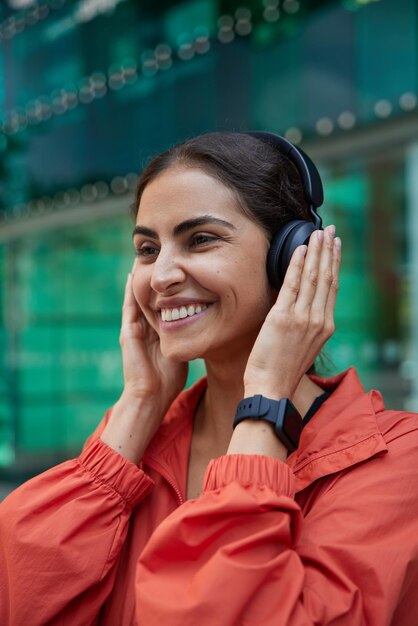 L'immagine verticale di una giovane donna positiva con i capelli scuri tiene le mani sulle cuffie stereo vestite con una giacca a vento e indossa un moderno smartwatch sorride felicemente posa all'aperto su sfondo sfocato