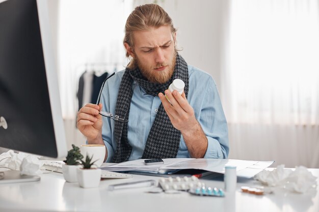 L'impiegato di concetto maschio barbuto malato in camicia e sciarpa blu con gli occhiali si è concentrato sulla lettura della prescrizione delle pillole. Giovane manager con influenza, siede sul posto di lavoro circondato da droghe, compresse, vitamine