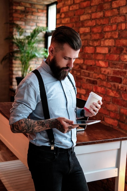 L'imponente uomo barbuto è in piedi accanto al tavolo. Indossa camicia e bretelle. Ha tatuaggi sulle braccia e sul collo. Sta tenendo un tablet e una tazza di caffè. L'uomo sta leggendo.