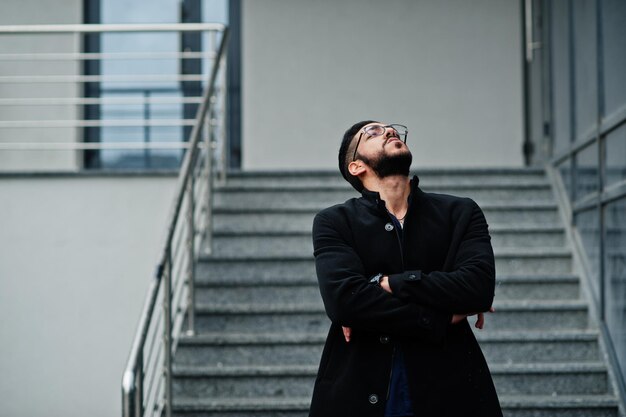 L'imprenditore mediorientale indossa un cappotto nero e occhiali da camicia blu contro il fumo di sigaretta dell'edificio per uffici
