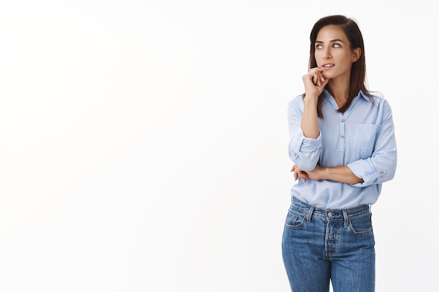 L'imprenditrice caucasica adulta premurosa e intrigante di bell'aspetto ha un pensiero interessante, guarda a sinistra interessato meditando, tocca il mento, cerca ispirazione, ha un'idea in mente, sta in piedi sul muro bianco