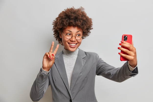 L'imprenditrice femminile positiva fa il gesto di pace prende selfie tramite smartphone gode di videoconferenza con il collega indossa abiti formali grigi in posa al coperto
