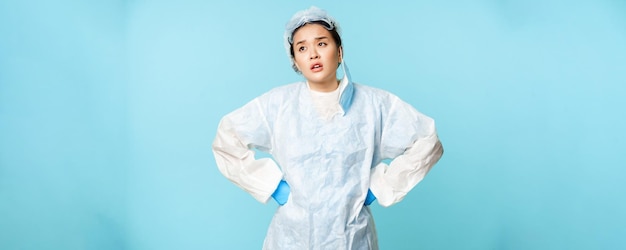 L'infermiere o il medico asiatico stanco si toglie la maschera medica sentendosi affaticato indossando dispositivi di protezione individuale sospirando con le mani sullo sfondo blu della vita