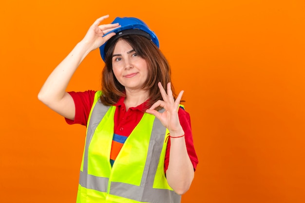 L'ingegnere della donna in maglia della costruzione e casco di sicurezza che sembrano il casco commovente di gesto di saluto sicuro che fa bene firma sopra la parete arancio isolata