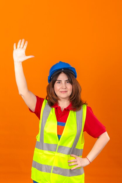 L'ingegnere della donna in maglia della costruzione e casco di sicurezza con il sorriso che ondeggia a qualcuno con consegna la parete arancio isolata
