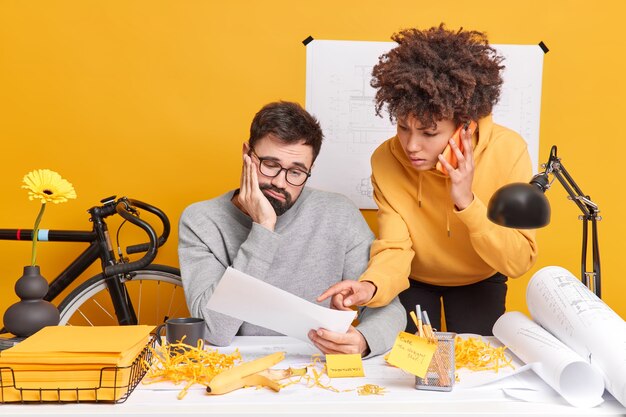 L'ingegnere maschio professionista guarda tristemente la carta ascolta la spiegazione della tirocinante che parla tramite la posa dello smartphone nello spazio di coworking insieme. Gli operai edili lavorano sui piani tecnici