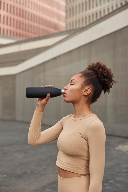 L'inquadratura laterale di una giovane donna sportiva con i capelli ricci pettinati beve acqua fresca ha sete dopo che l'allenamento cardio conduce uno stile di vita sano vestito con abbigliamento sportivo Concetto di sport e benessere delle persone