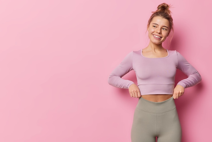 Foto gratuito l'inquadratura orizzontale di una donna sportiva felice vestita con una tuta da ginnastica ha un corpo sano in forma distoglie lo sguardo ha pose di espressione allegra su sfondo rosa con spazio per la copia per i tuoi contenuti pubblicitari.