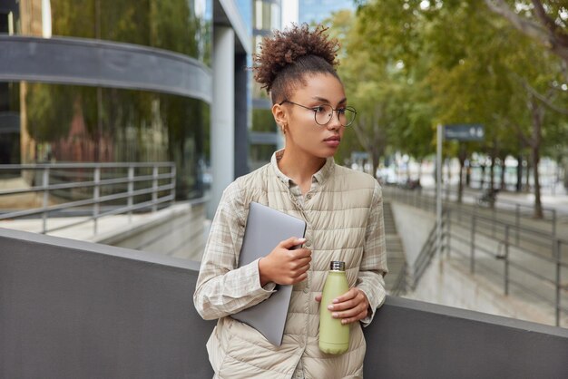 L'inquadratura orizzontale di una giovane donna dai capelli ricci premurosa indossa una camicia e un gilet tiene una moderna tavoletta digitale e una bottiglia di acqua fresca focalizzata lontano posa all'aperto su sfondo urbano sfocato