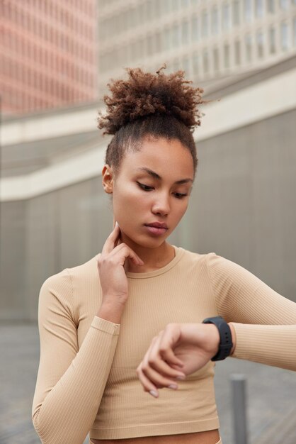 L'inquadratura verticale di una seria donna dai capelli ricci controlla il polso sul collo focalizzato sullo smartwatch vestito con abbigliamento sportivo monitora la frequenza cardiaca dalle pose del polso all'aperto La donna sportiva utilizza il tracker di allenamento