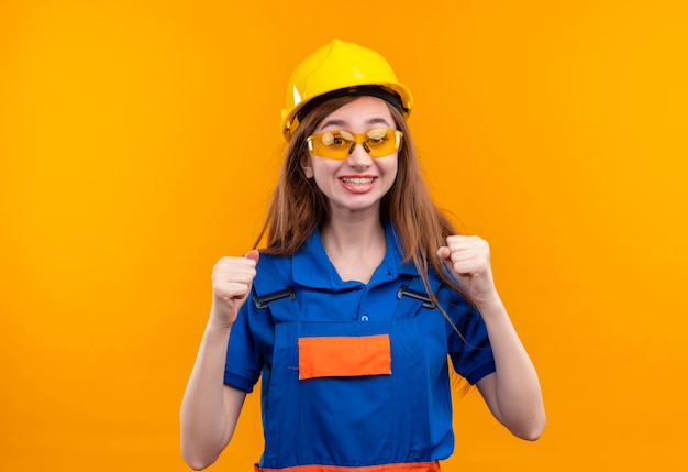 L'operaio del costruttore della giovane donna in uniforme della costruzione e il casco di sicurezza aumentano i pugni usciti e gioiscono felice del suo successo che sta sopra la parete arancione