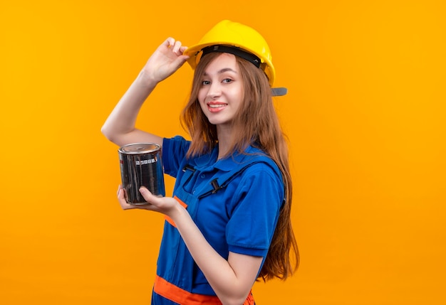 L'operaio del costruttore della giovane donna in uniforme della costruzione e la vernice della tenuta del casco di sicurezza può guardare fiducioso toccando il suo casco che sta sopra la parete arancione