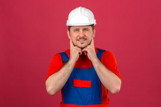 L'uniforme d'uso della costruzione dell'uomo e il casco di sicurezza del costruttore tengono il dito sulle guance che sorridono controllando la parete rosa isolata