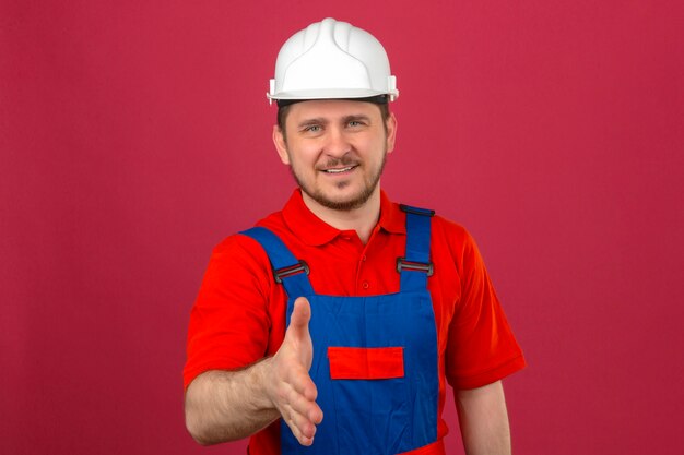 L'uniforme della costruzione dell'uomo del costruttore e il casco di sicurezza che sorridono facendo gesto di saluto amichevole che offrono la mano che controllano la parete rosa isolata