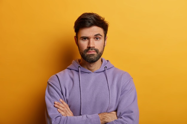 L'uomo adulto barbuto dall'aspetto serio tiene le mani incrociate, sembra assertivo, alza le sopracciglia con meraviglia, giudica la presentazione del collega, indossa una felpa con cappuccio, isolato su un muro giallo