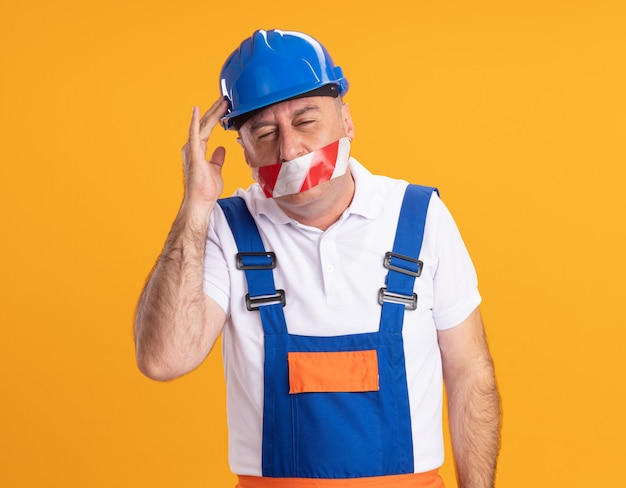 L'uomo adulto caucasico dispiaciuto del costruttore in uniforme copre la bocca con del nastro adesivo e mette la mano sulla testa sull'arancio
