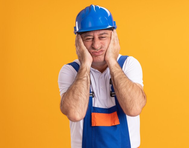 L'uomo adulto caucasico infastidito del costruttore in uniforme mette le mani sulla faccia sull'arancia