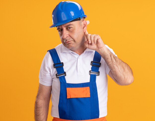 L'uomo adulto caucasico senza tracce del costruttore in uniforme tiene il dito dietro l'orecchio sull'arancia