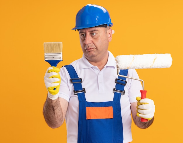 L'uomo adulto confuso del costruttore in guanti protettivi da portare dell'uniforme tiene il pennello e la spazzola del rullo isolati sulla parete arancione