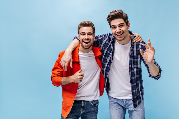 L'uomo affascinante in giacca arancione mostra il pollice in su Ragazzo Brunet in camicia a scacchi e il suo amico sorride su sfondo blu