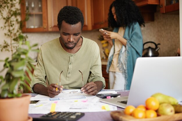 L'uomo africano con gli occhiali e la matita nelle sue mani guardando con frustrazione i documenti davanti a lui mentre fa il lavoro di ufficio, cercando di pagare tutti i debiti familiari, seduto al tavolo con laptop e calcolatrice