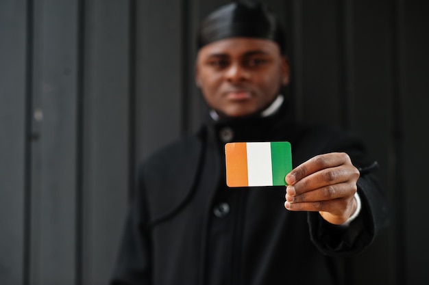 L'uomo africano indossa il durag nero tiene la bandiera della Costa d'Avorio a portata di mano isolata su sfondo scuro