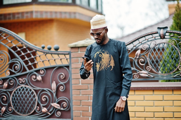 L'uomo africano ricco in abiti tradizionali alla moda e cappello ha posato sullo sfondo all'aperto la sua villa parlando sul telefono cellulare