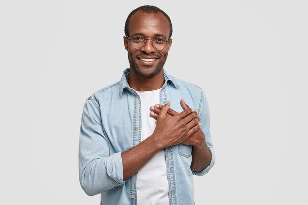 L'uomo africano toccato e grato tiene le mani sul petto, esprime gratitudine, guarda piacevolmente la telecamera