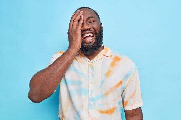 L'uomo afroamericano barbuto allegro ottimista fa ridere il palmo della faccia esprime felicemente emozioni autentiche felici vestito con una maglietta sbiadita isolata sul muro blu