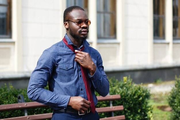 L&#39;uomo afroamericano bello ripara il suo legame rosso sulla camicia blu che sta fuori