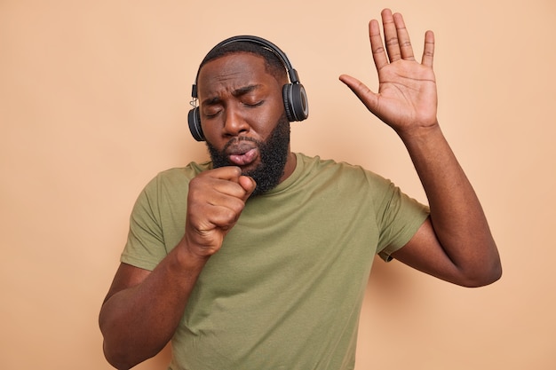 L'uomo afroamericano canta la canzone tiene la mano vicino alla bocca come se il microfono ascoltasse musica dal giocatore usa le cuffie wireless indossa una maglietta casual si diverte durante il tempo libero isolato sul muro beige