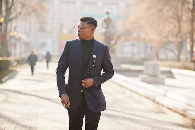 L'uomo afroamericano dall'aspetto sorprendente indossa un blazer blu con spilla a collo alto nero e occhiali posati in strada Ragazzo nero alla moda