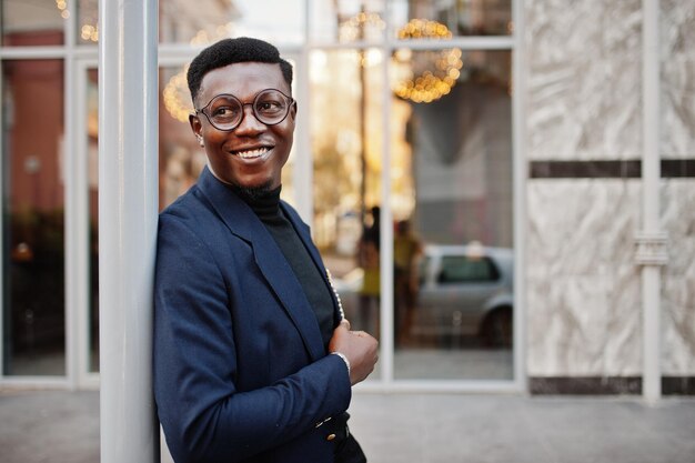 L'uomo afroamericano dall'aspetto sorprendente indossa un blazer blu con spilla a collo alto nero e occhiali posati in strada Ragazzo nero alla moda