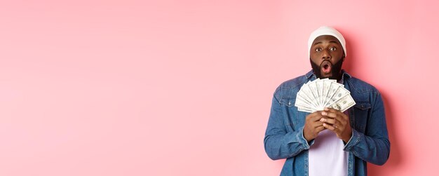 L'uomo afroamericano stupito riceve un premio in denaro mostrando denaro e fissando la telecamera con soggezione in piedi ov