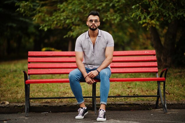 L'uomo alla moda con barba araba alta indossa su camicia jeans e occhiali da sole seduto su una panchina rossa al parco